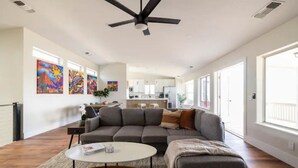 A spacious living room with a gray sectional sofa, a round coffee table, and a ceiling fan. There are large windows on one side and a kitchen in the background.
