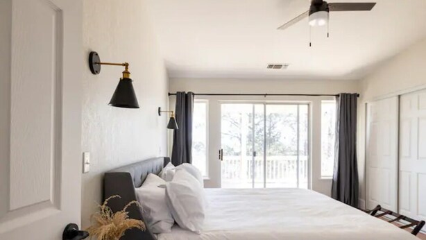 A bedroom with a bed, a nightstand, a wall-mounted lamp, a ceiling fan, and a view of the outdoors through a sliding door.