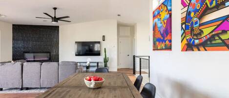 Ein modernes Wohnzimmer mit einem Holzesstisch, grauen Sofas, einem Flachbildfernseher und farbenfroher Kunst an den Wänden.