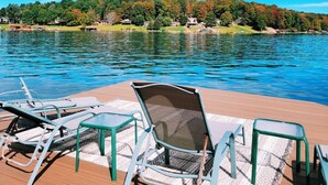 Una terraza de madera con sillones de descanso y una mesa, con vista a un lago y una orilla boscosa.