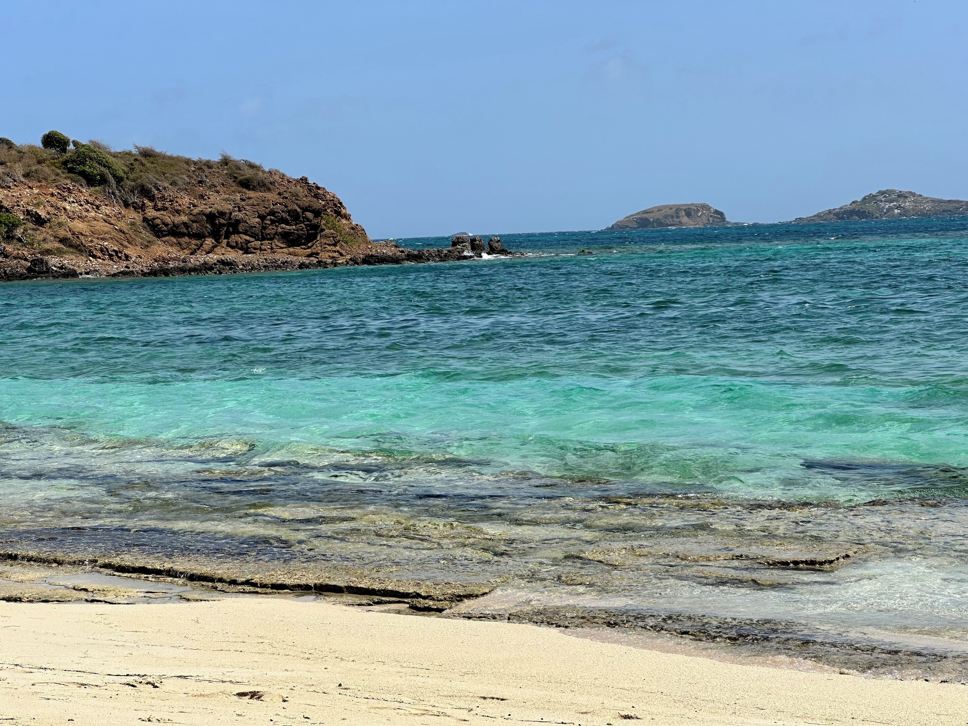 Una playa con agua turquesa cristalina, acantilados rocosos y islas a lo lejos.