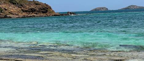 Una playa con agua turquesa cristalina, acantilados rocosos y islas a lo lejos.