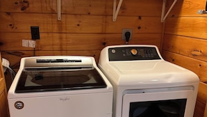 A laundry room with two white appliances, a dryer and a washer, and a wooden shelf above.