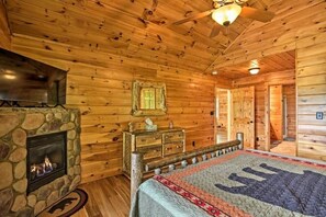 Interior de una cabaña de madera con chimenea de piedra, una cama con colcha estampada, un tocador de madera y un ventilador de techo.