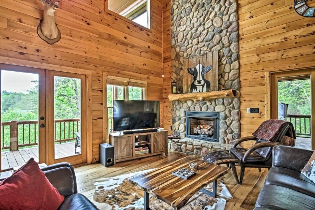 A wooden cabin interior with a stone fireplace, a television, and a deck.