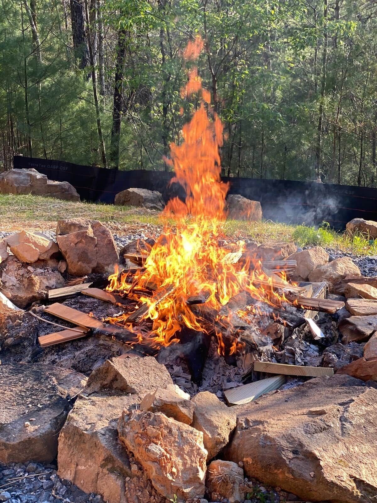 Ein Lagerfeuer, umgeben von Steinen und Baumstämmen in einem bewaldeten Gebiet.