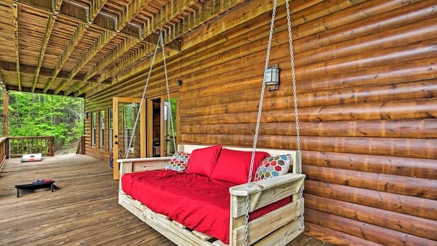 A wooden log cabin with a swing bed featuring a red blanket, a wooden deck, and a view of a forested area.