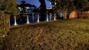 Vista nocturna de un puerto deportivo con barcos amarrados, un área verde en primer plano y un edificio con ventanas iluminadas al fondo.
