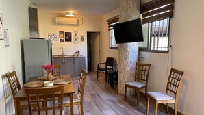 A dining area with a wooden table and chairs, a kitchen in the background, and a flat-screen TV mounted on a pillar. - Chalet in Batroun, 1BR, gazon, 65sqm, parking, wifi (محافظة الشمال)