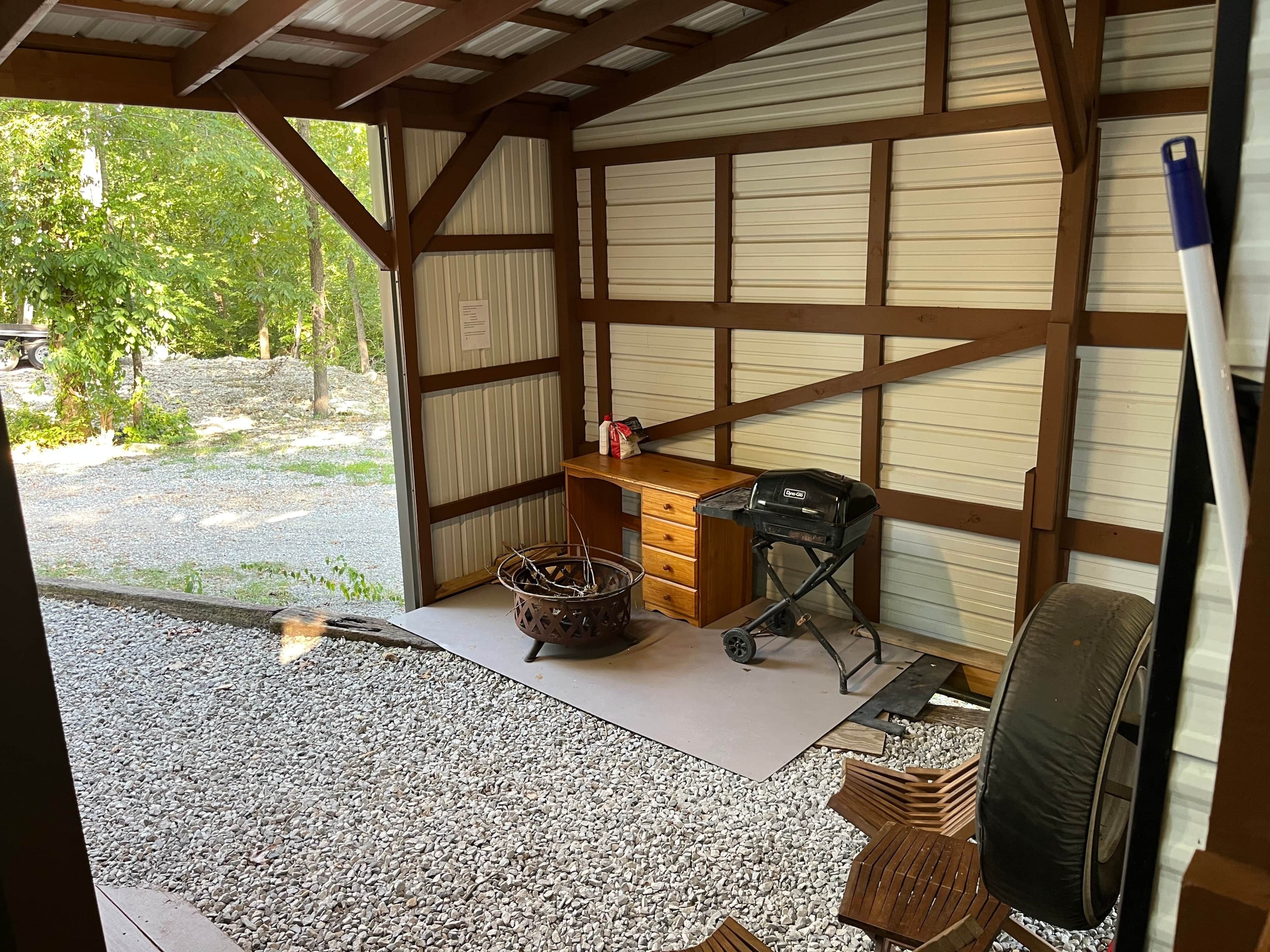 Eine Garage mit Dach, einem Kiesboden, einer Holzbank, einem Grill und einem Korb.