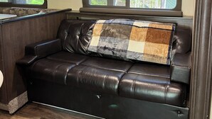 Un canapé en cuir noir agrémenté d’un coussin à motifs écossais, une fenêtre donnant sur des arbres et un plancher en bois.