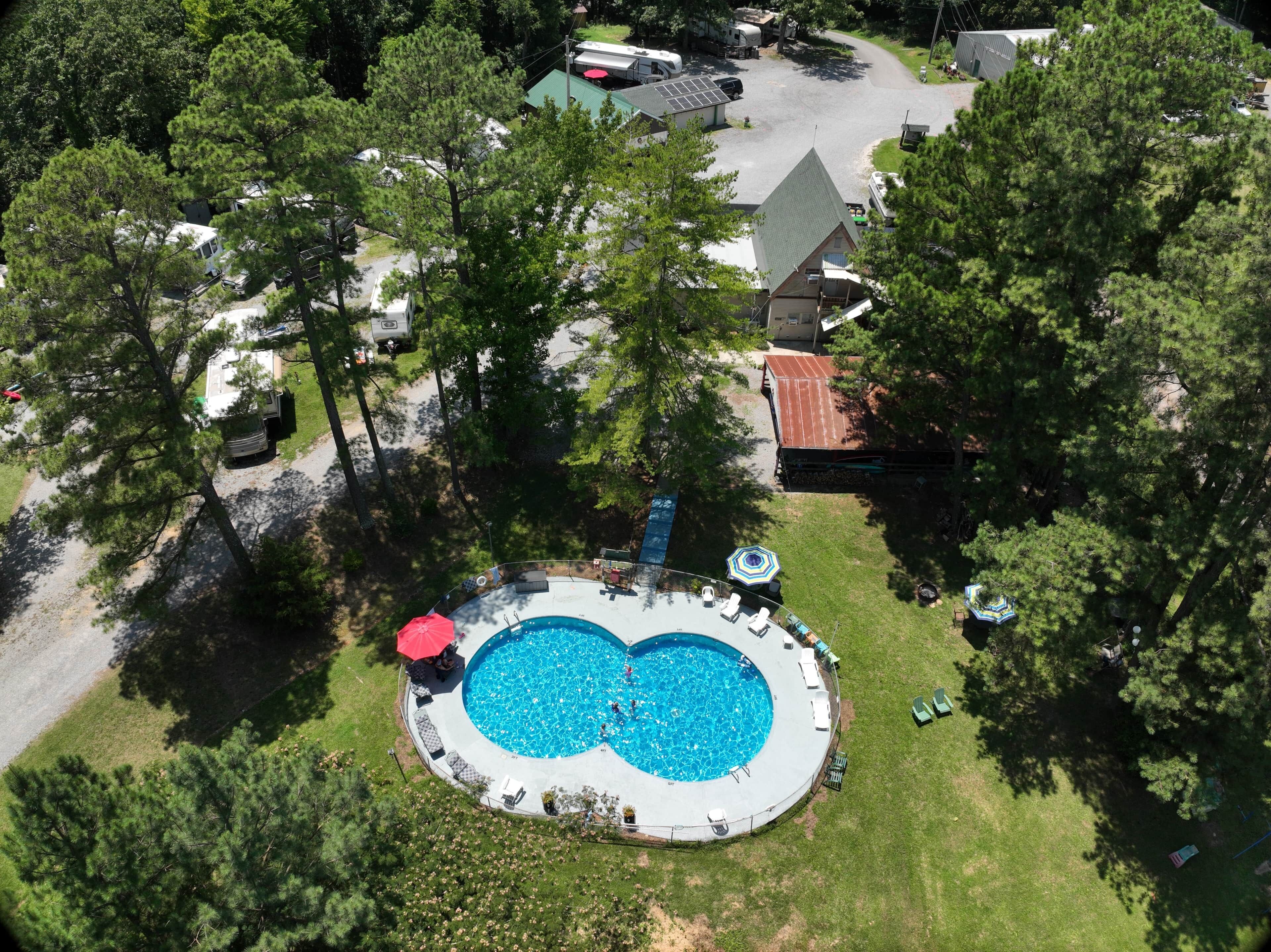 An aerial view of a property with a circular swimming pool surrounded by lounge chairs and umbrellas, a covered patio area, and a parking lot with RVs.
