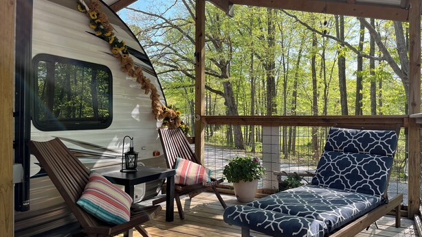 Un espace extérieur abrité doté d’une terrasse en bois, d’un camping-car blanc et d’un banc agrémenté d’un coussin à motifs.
