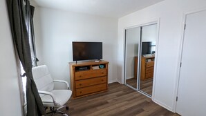 A room with a wooden dresser, a television on top, a white chair, and a sliding door with a mirror. - Adorable Townhome (Midland)