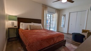A bedroom with a large bed, a nightstand with a lamp, a window with curtains, a door, and a chair. - Adorable Townhome (Midland)