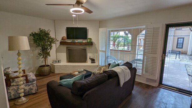 A living room with a fireplace, a ceiling fan, a TV mounted above the fireplace, and a large window with blinds. - Adorable Townhome (Midland)