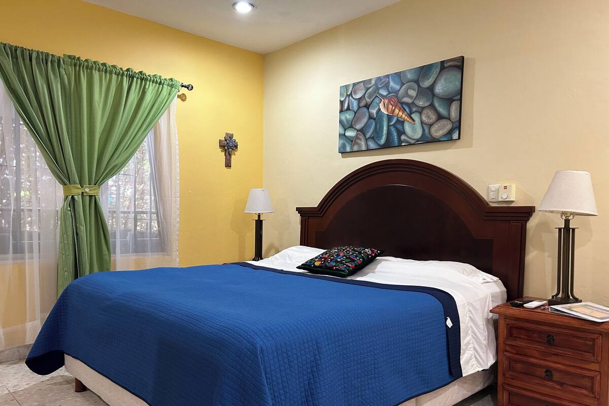 A neatly made bed with a blue bedspread, a green curtain, a wall clock, a painting of a shell, and a bedside table with a lamp.