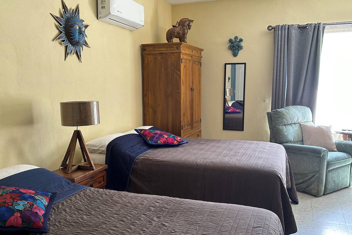 A hotel room with two beds, a wooden wardrobe, a mirror, a decorative sun, and a window with curtains.