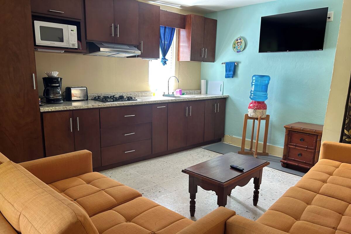 A compact living space with a kitchenette, a sofa set, a small table, and a water dispenser.
