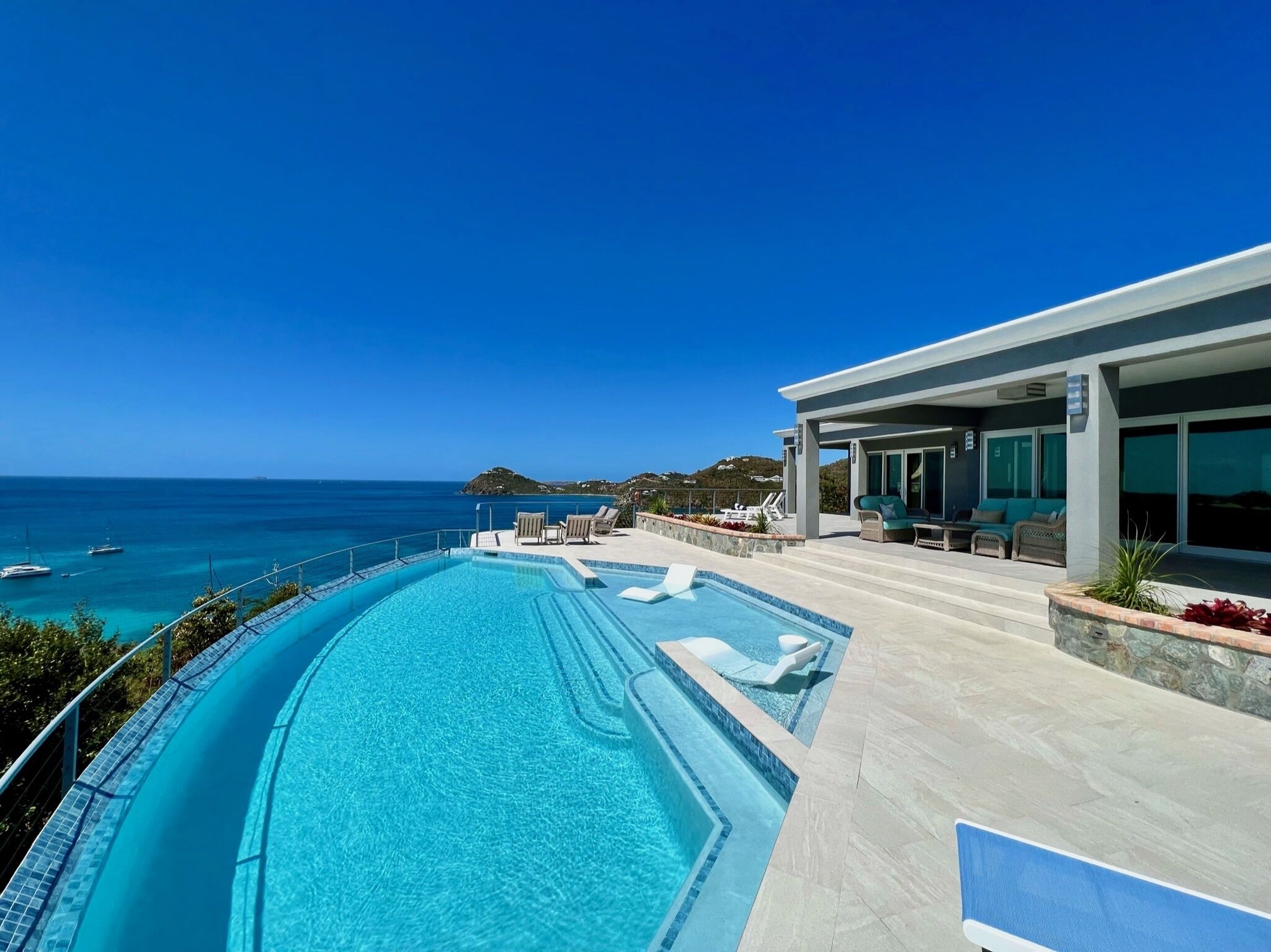 Ein moderner Poolbereich mit klarem, blauem Wasser, Liegestühlen und Blick auf das Meer und die Hügel im Hintergrund.