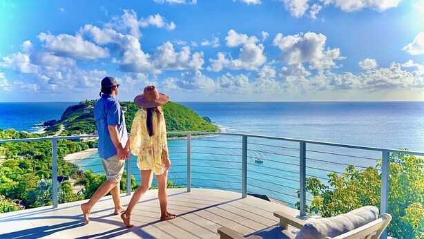 Una pareja caminando sobre una terraza con vistas a la costa, cielo despejado y una isla distante.