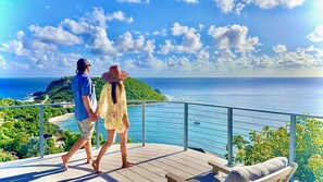 Un couple se promène sur une terrasse qui offre une vue sur la côte, avec un ciel dégagé et une île visible au loin.