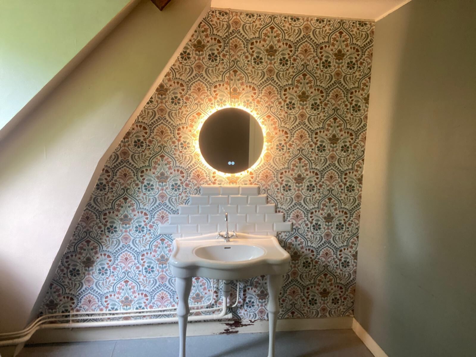 A bathroom with a patterned tile wall, a white sink, and a round mirror with a light above it.