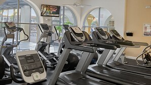 Une salle de sport équipée de plusieurs tapis de course et de vélos d’exercice, de grandes fenêtres à arc en plein cintre et d’un ventilateur de plafond.