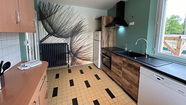 A compact kitchen with a tiled floor, wooden cabinets, a black range hood, a built-in dishwasher, and a window with a view of greenery outside.