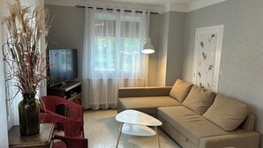 A living room with a beige sofa, a small white table, a red wicker chair, a TV, and a vase with dried plants.
