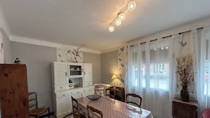 A dining area with a table covered in a polka dot tablecloth, surrounded by wooden chairs. A white cabinet with glass doors is against the wall, and a lamp is on a small table beside the window.