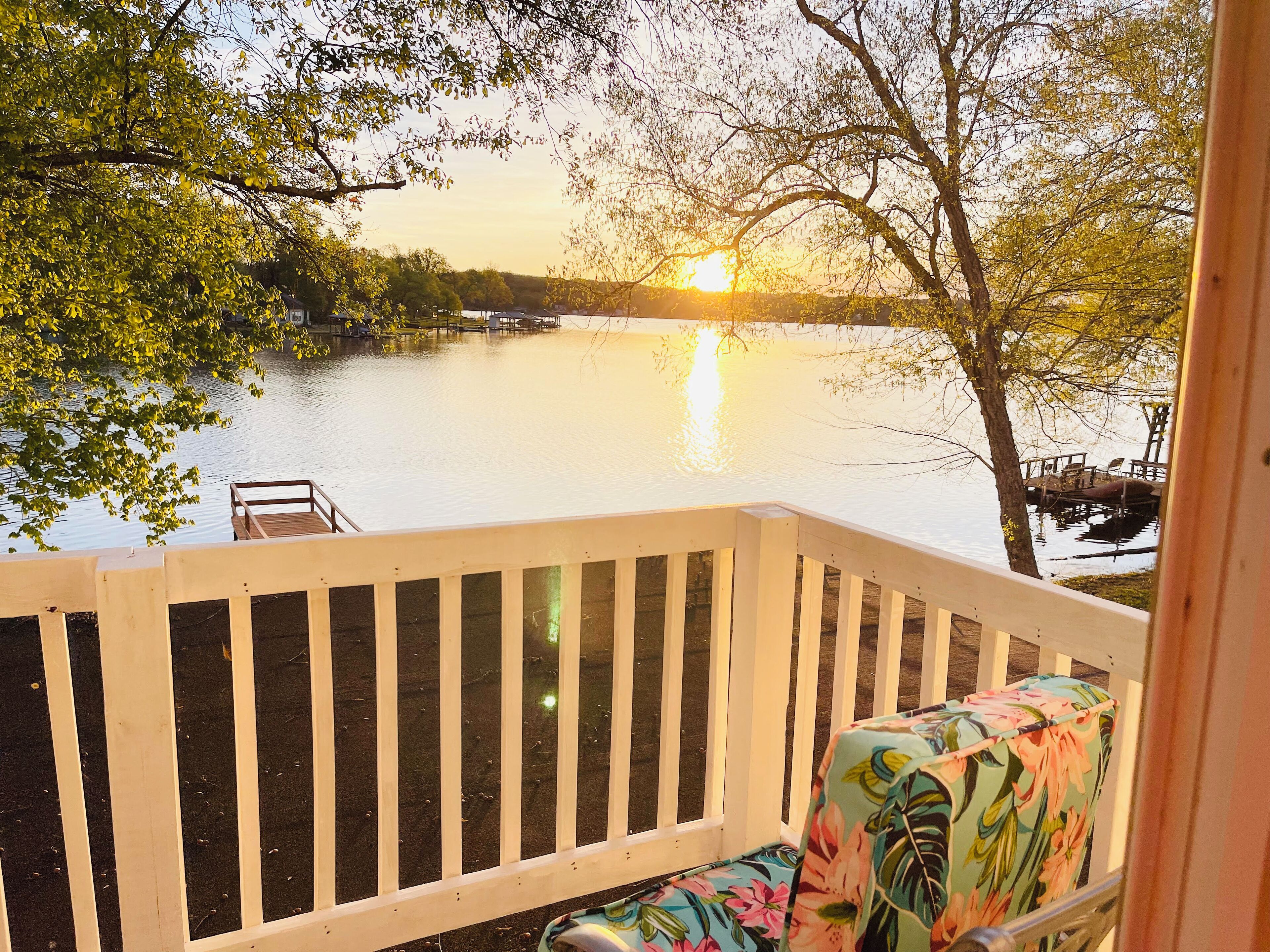 A wooden deck with a white railing overlooking a calm lake at sunset.