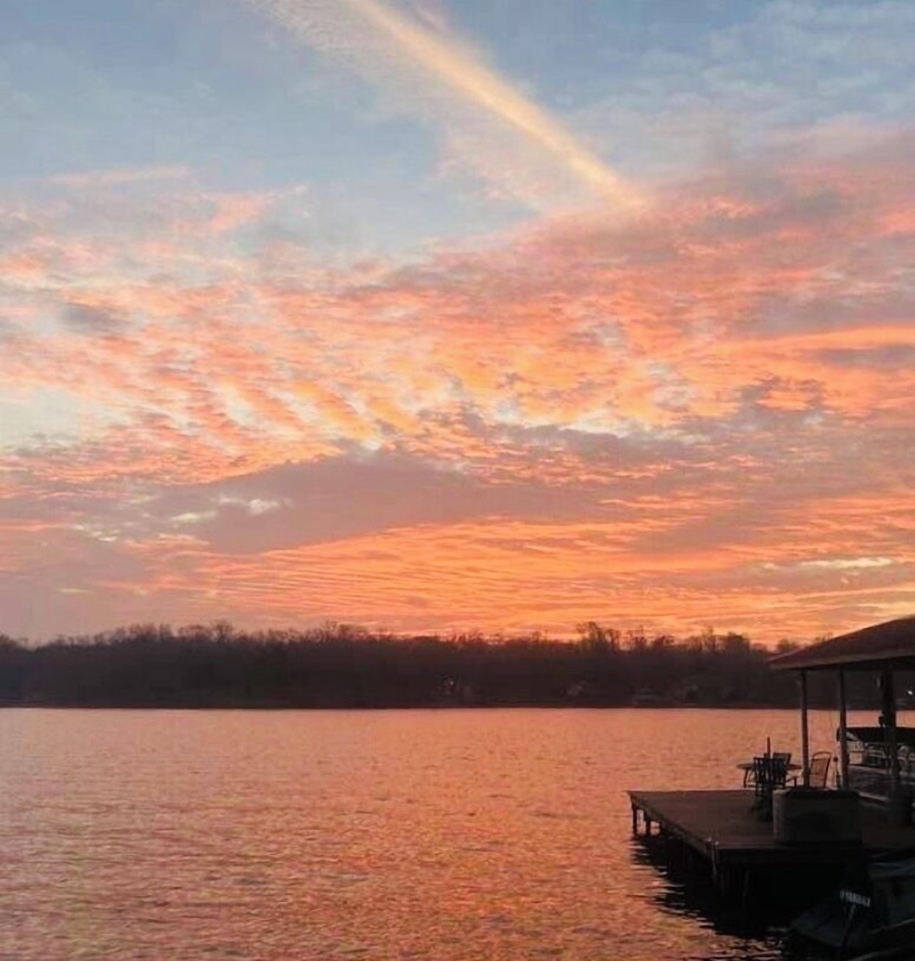 A sunset over a calm lake with a dock and chairs.