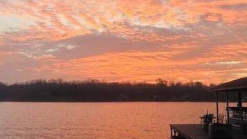 A sunset over a calm lake with a dock and chairs.