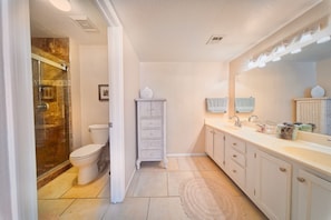 A bathroom with a glass shower enclosure, a toilet, a wicker chest of drawers, a sink with a mirror, and a lighted vanity.