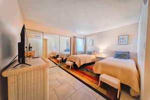 A hotel room with two beds, a television, a wicker side table, and a colorful rug.