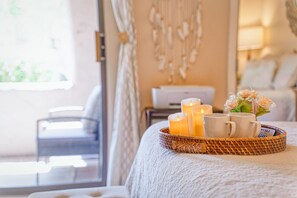 A cozy bedroom with a bed, a bedside table with candles, and a wicker tray with cups and flowers.