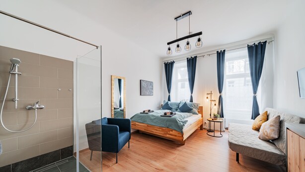 A modern bedroom with a glass shower, a bed with a wooden frame, a sofa, and a small side table. - 3 Bedrooms - 75sqm - 15 Min. City Center (Vienna)