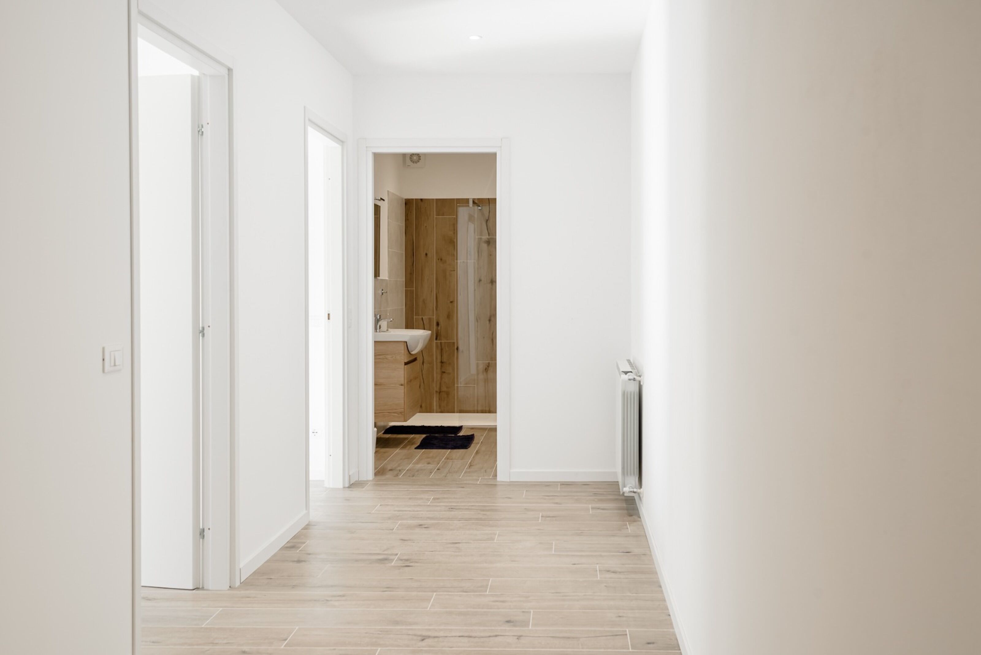 Un couloir doté d’un plancher en bois, de murs blancs et d’une salle de bain visible, avec une porte en bois et un lavabo.