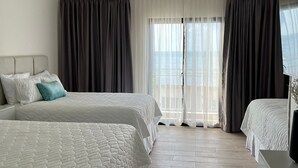 Habitación de hotel con dos camas, ventilador de techo, televisor y vistas al mar a través de amplias ventanas.