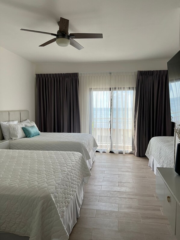A hotel room with two beds, a ceiling fan, a TV, and a view of the sea through large windows.