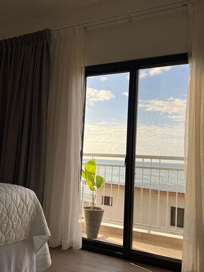 A hotel room with a bed, a potted plant, and a balcony with a view of the sea.
