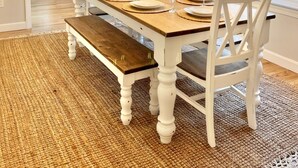 Un comedor con una mesa de madera dispuesta para cuatro personas, sillas blancas y una alfombra tejida.