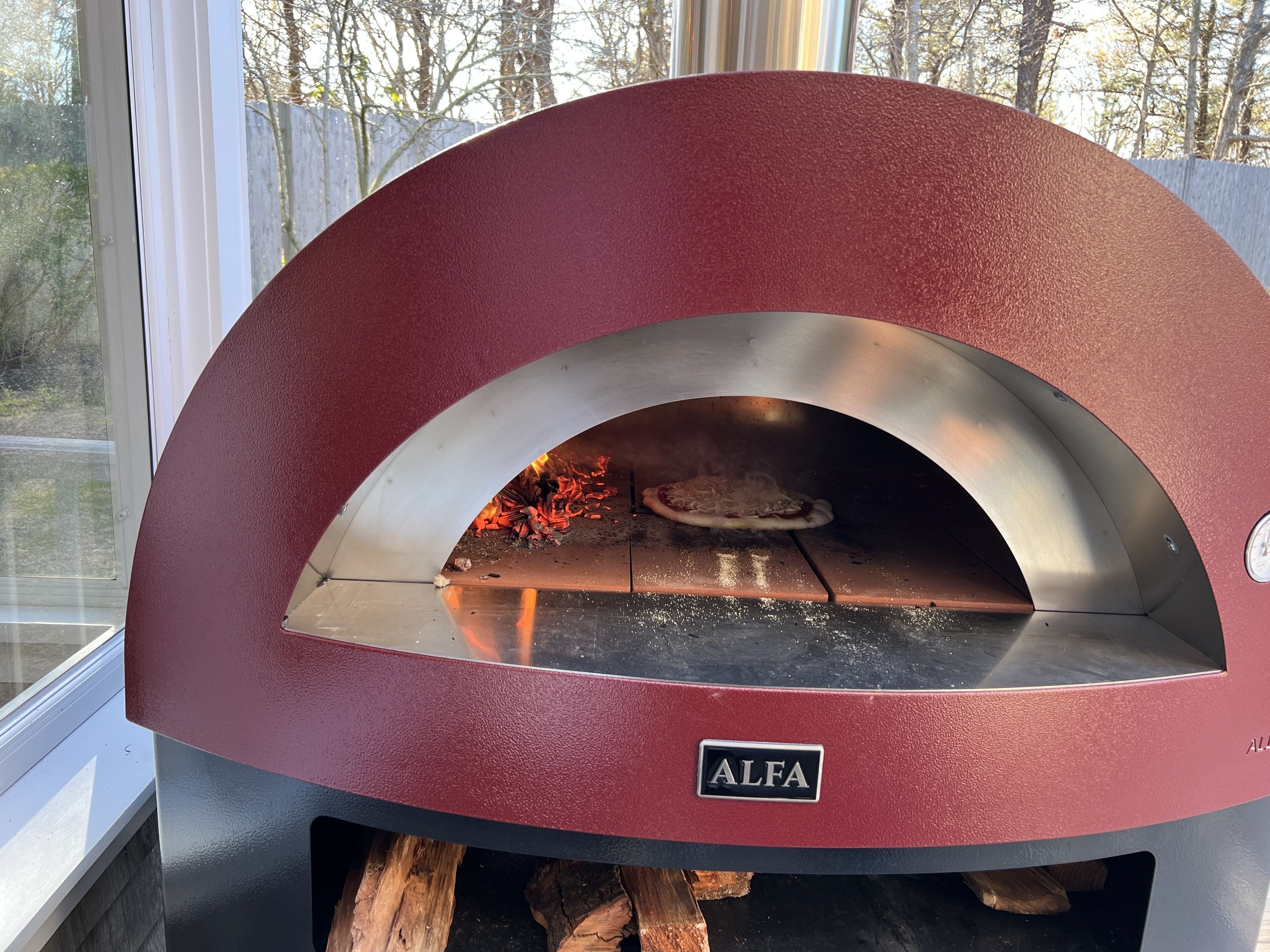 Punainen ALFA-merkkinen puu-uunipizzauuni, jossa on lasinen etuosa, minkä ansiosta uunin sisällä oleva tuli ja pizza ovat näkyvissä.