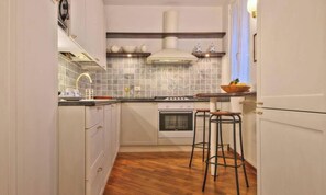 A narrow kitchen with white cabinets, a tiled backsplash, a stove, and two bar stools.