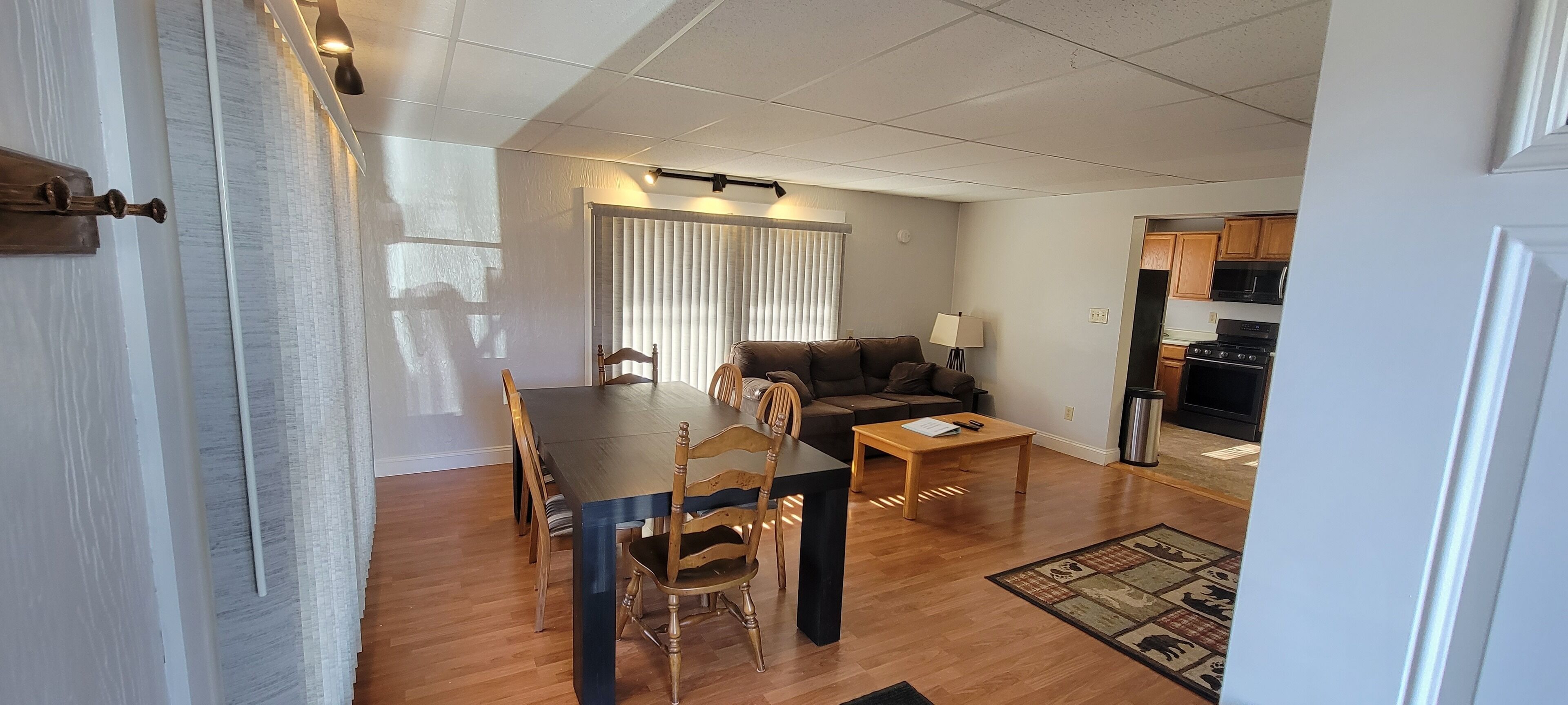 A living room with a dining table, chairs, a sofa, and a coffee table. There is a kitchen area with cabinets and appliances visible.