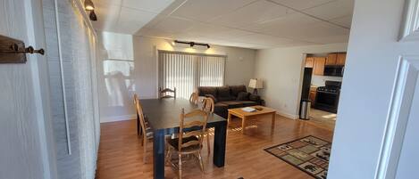 A living room with a dining table, chairs, a sofa, and a coffee table. There is a kitchen area with cabinets and appliances visible.