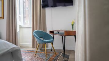 A room with a desk, a blue chair, a TV mounted on the wall, and a window with curtains.