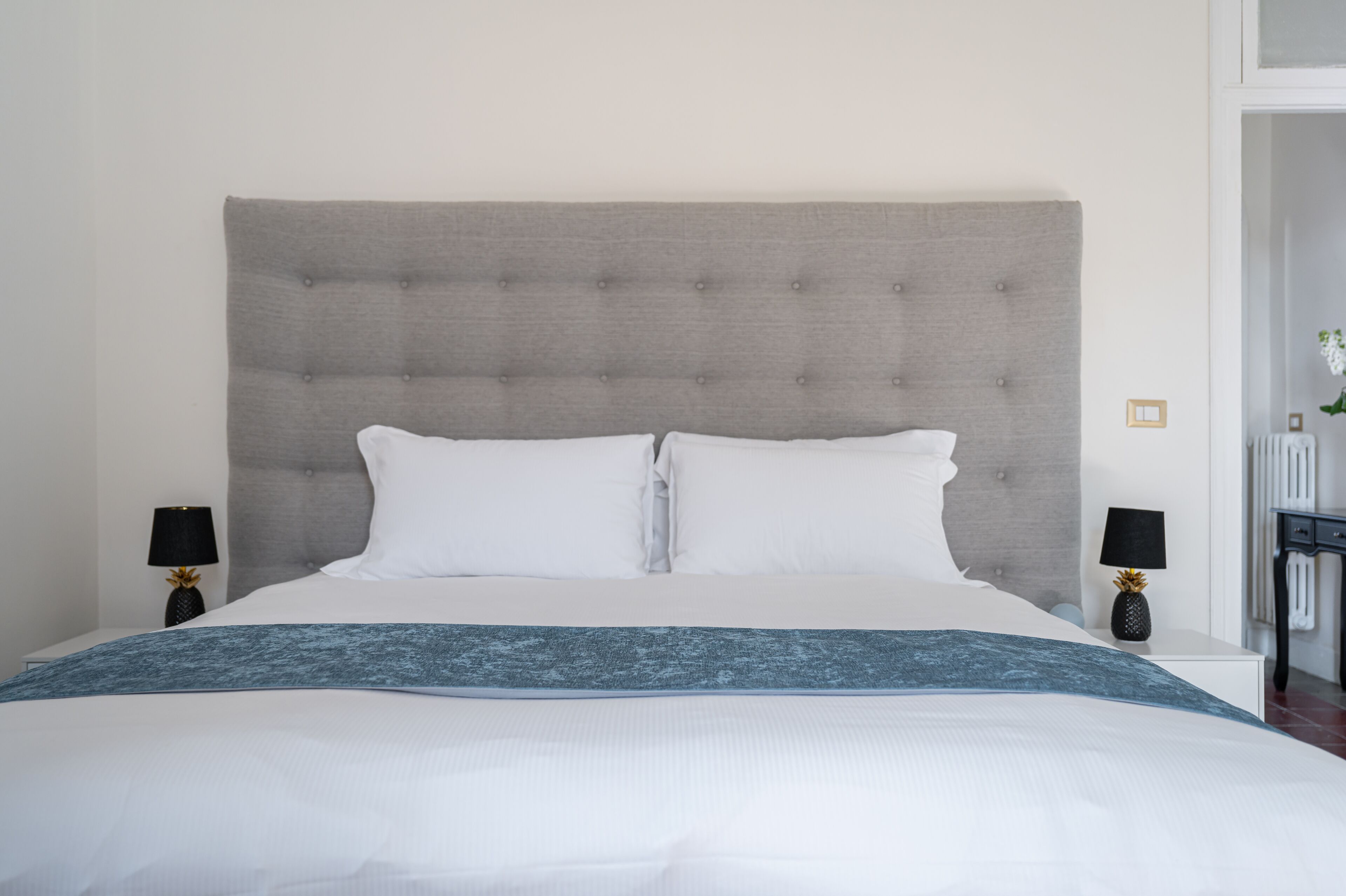 A neatly made bed with a grey upholstered headboard, white pillows, and a blue throw blanket. There are two bedside tables with lamps and decorative items.