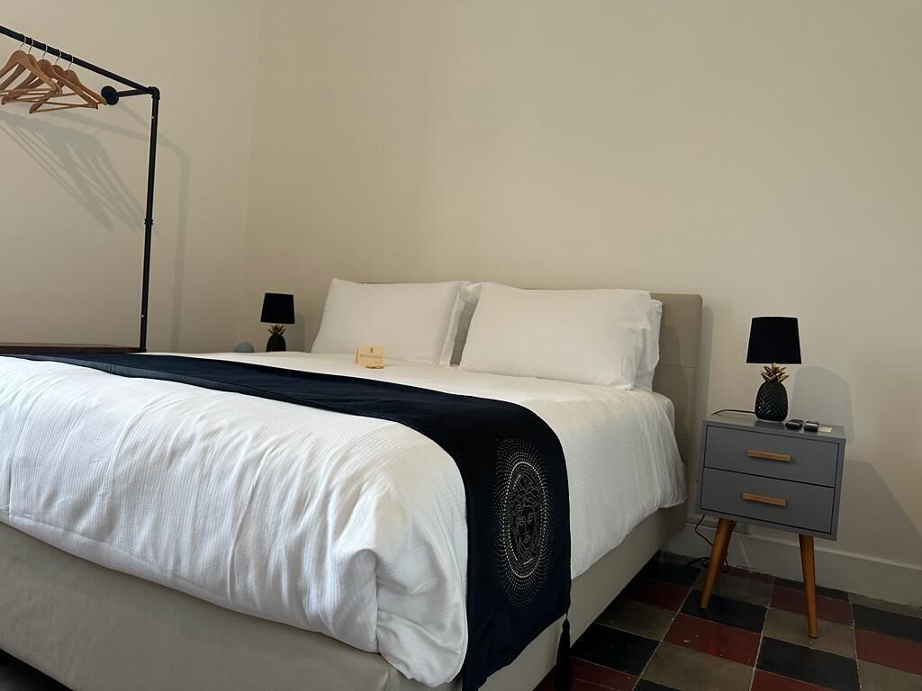 A neatly made bed with white linens and a dark throw blanket, a bedside table with a lamp, and a wall-mounted clothing rack.